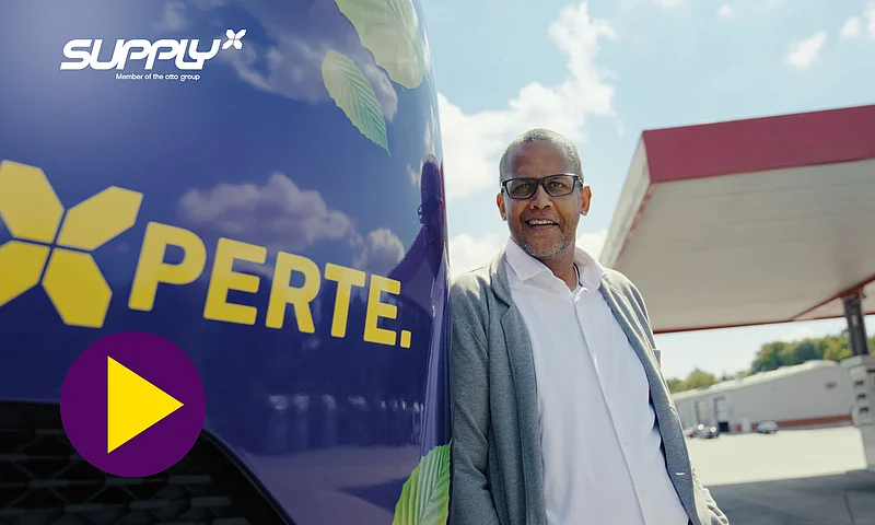 Ein lächelnder Mann lehnt an einem lila SupplyX-LKW mit der Aufschrift "XPERTE." und einem Play-Button-Symbol, was auf das YouTube-Video "SupplyX is hitting the road for the environment" über nachhaltige Logistik hinweist.