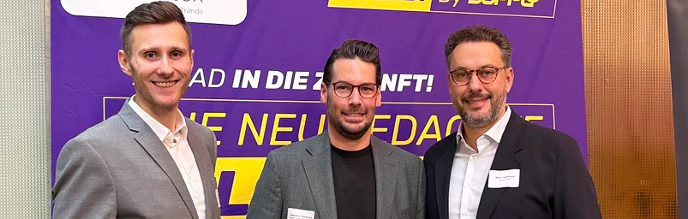 Stephan Pfeifer (Kontakt für AHEAD. By SupplyX), Marco Hoffmeyrer (CEO scoretex) und ein weiterer SupplyX-Vertreter lächeln auf dem BTE-Kongress 2025 vor einem Banner, das Logistik als Gamechanger im Modehandel hervorhebt.
