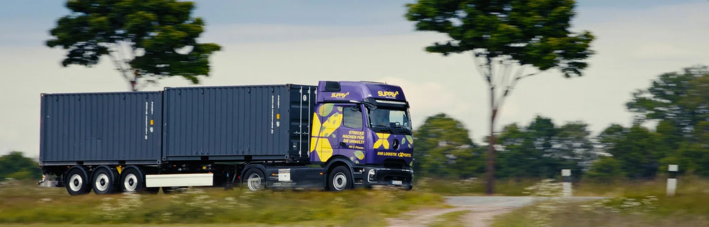 Ein lila SupplyX E-LKW mit Containern fährt auf einer ländlichen Straße.