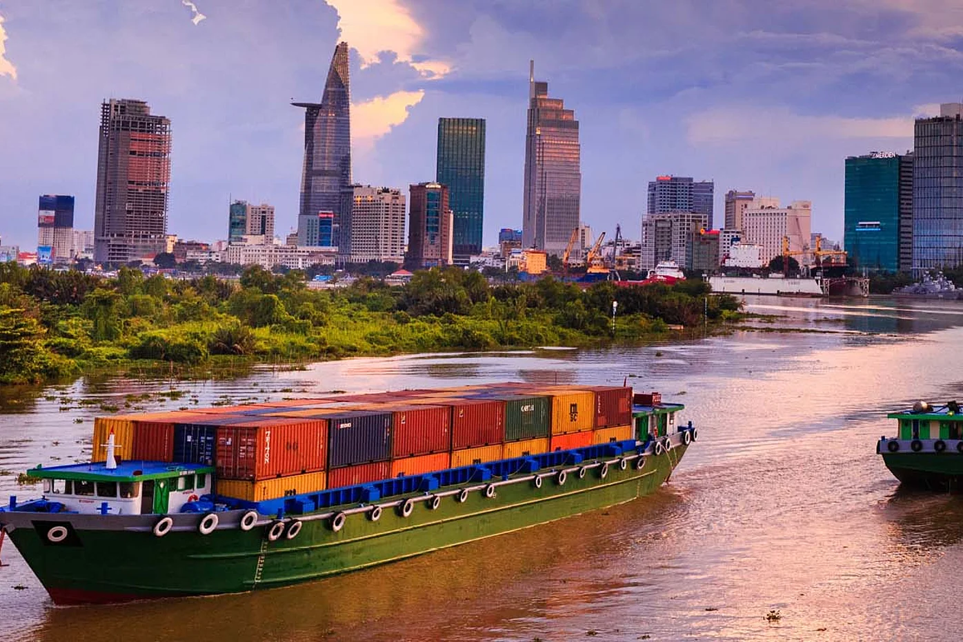 Containerschiffe auf einem Fluss vor der Skyline einer vietnamesischen Stadt – Symbol für Chancen und Risiken des Produktionsstandorts Vietnam in globalen Lieferketten.