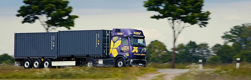 Ein lila SupplyX E-LKW mit Containern fährt auf einer ländlichen Straße.