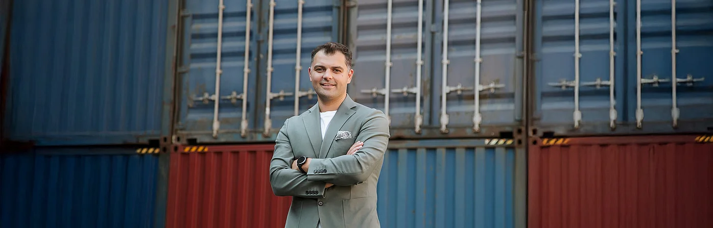 Porträt von Robert Klukowski, dem neuen Leiter der SupplyX-Niederlassung in Gdynia, der lächelt und die Arme verschränkt vor gestapelten Containern steht.