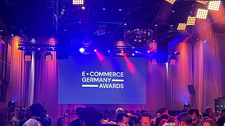 Blick in einen festlich beleuchteten Veranstaltungsraum mit vielen Gästen, im Hintergrund eine Bühne mit einer großen Leinwand, auf der "E-COMMERCE GERMANY AWARDS" zu lesen ist.